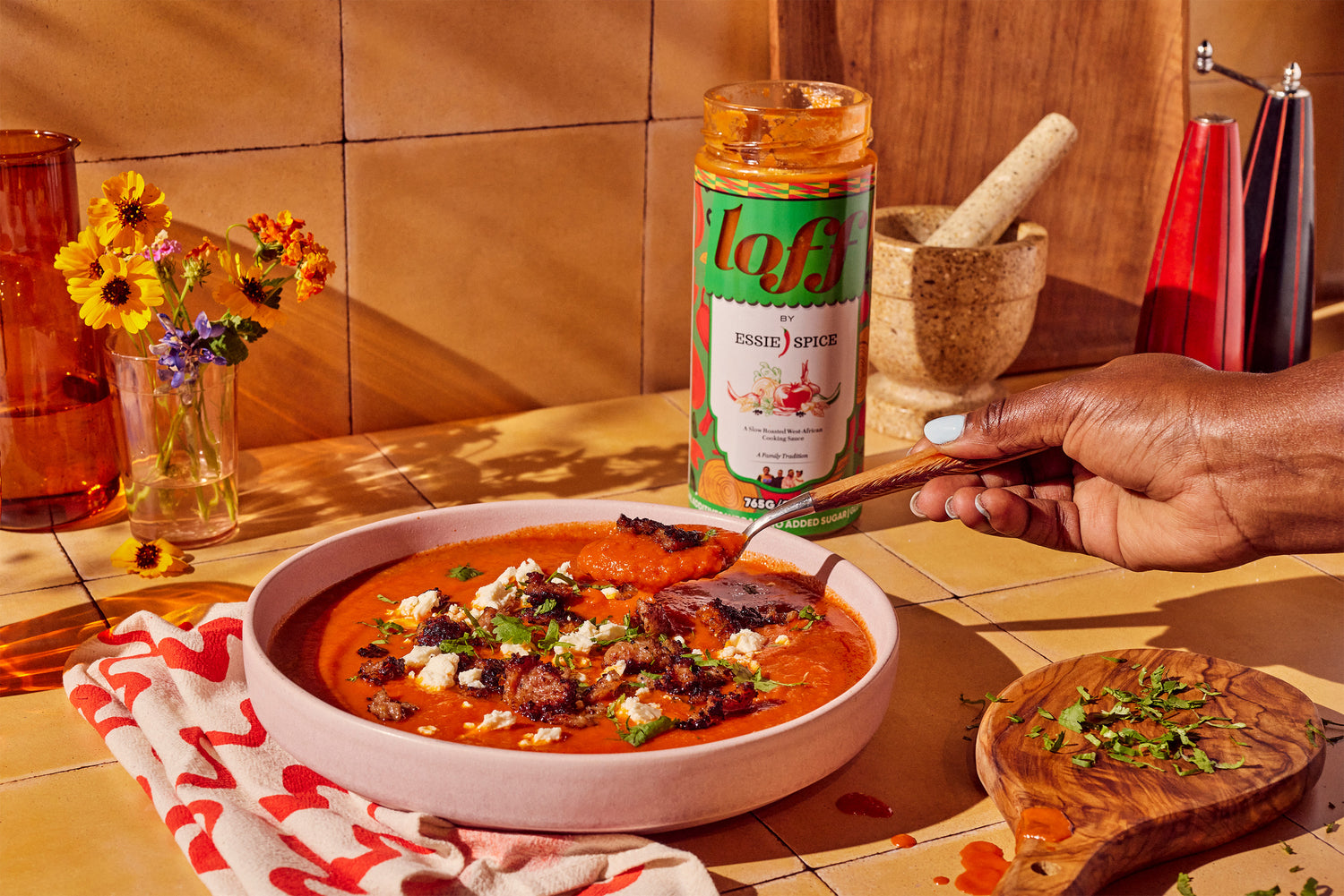 A hand adding sauce to a bowl of soup with a jar of spice, a mortar and pestle, flowers, and herbs on a table.